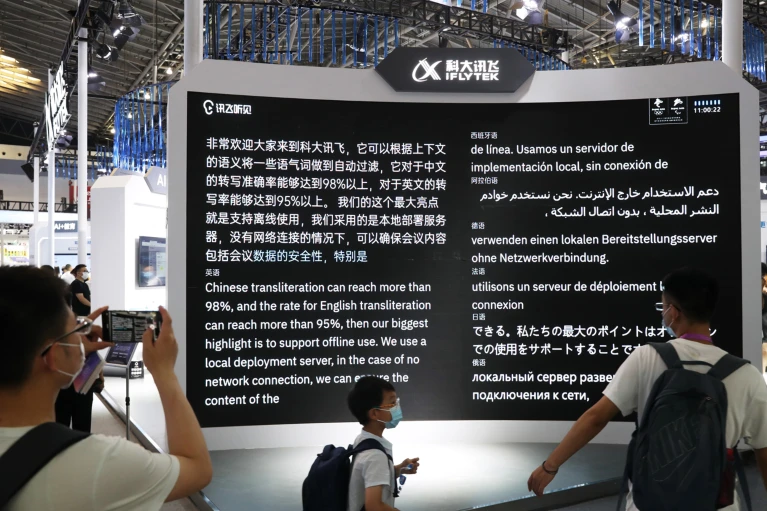 A group of people at a tech exhibition observe a large display screen from iFlytek. The screen showcases multilingual text about Chinese and English transliteration capabilities, emphasizing offline use and local deployment without network connection. The text is presented in multiple languages, including Chinese, English, Spanish, Arabic, German, French, Japanese, and Russian. One person in the foreground is taking a photo or video of the screen with a tablet, while others are standing and reading the information.